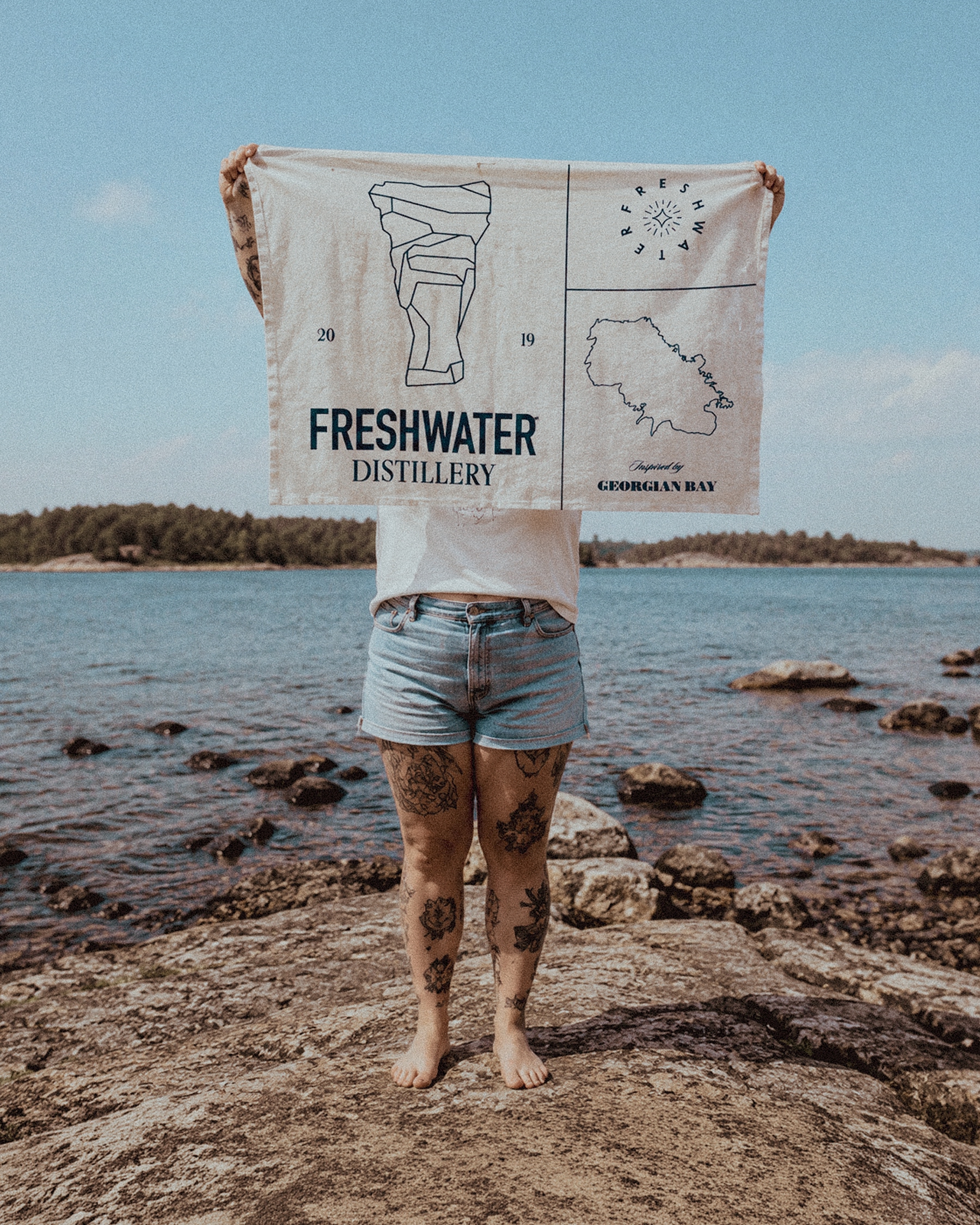Person standing on the rocky Georgian Bay shoreline displaying a Freshwater Distillery flag.